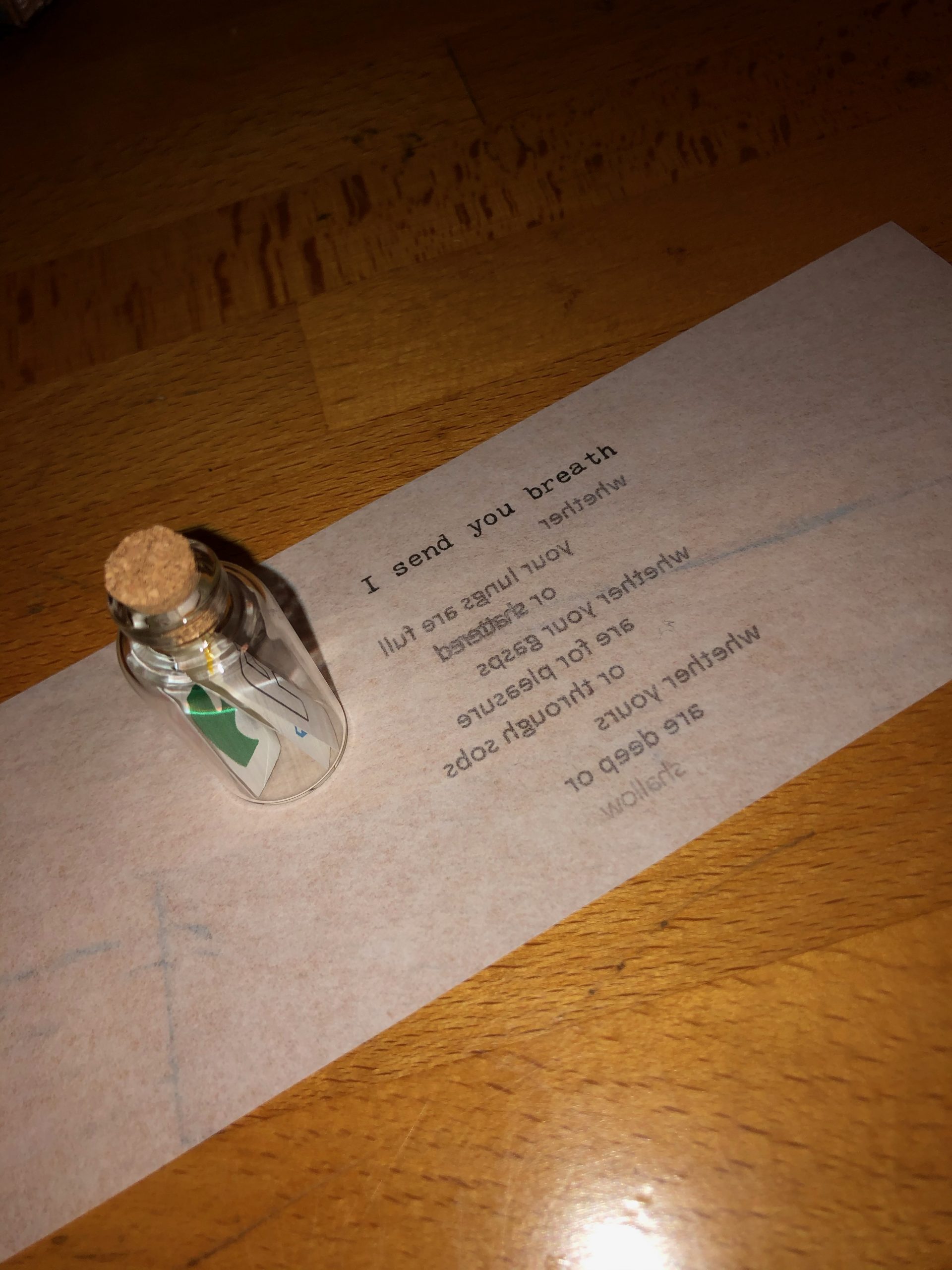 Image of a miniature bottle with a note inside, and a strip of transparent paper on a wooden surface. The transparency is upside down, so the words read backwards: “I send you breath/ whether/ your lungs are full/ or shattered/ whether your gasps/ are for pleasure/ or through sobs/ whether yours/ are deep or/ shallow”.